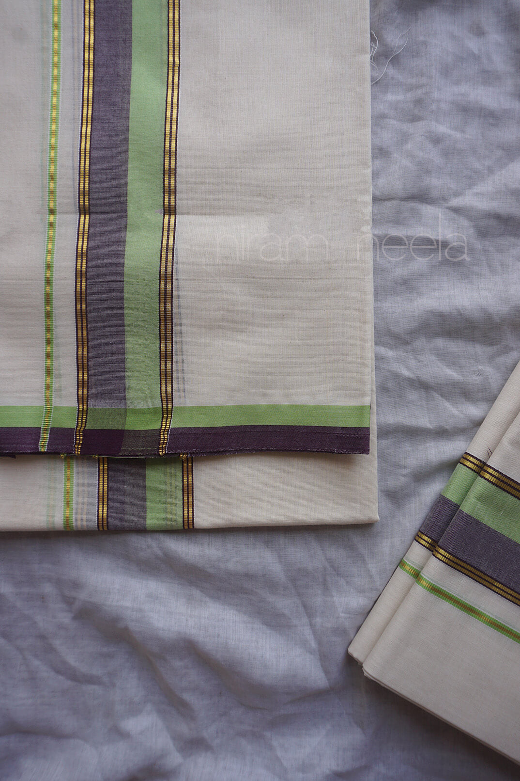 A folded cream-colored cotton set mundu, featuring vertical stripes in lime green, dark purple, and gold, laid out on a textured light gray fabric background.