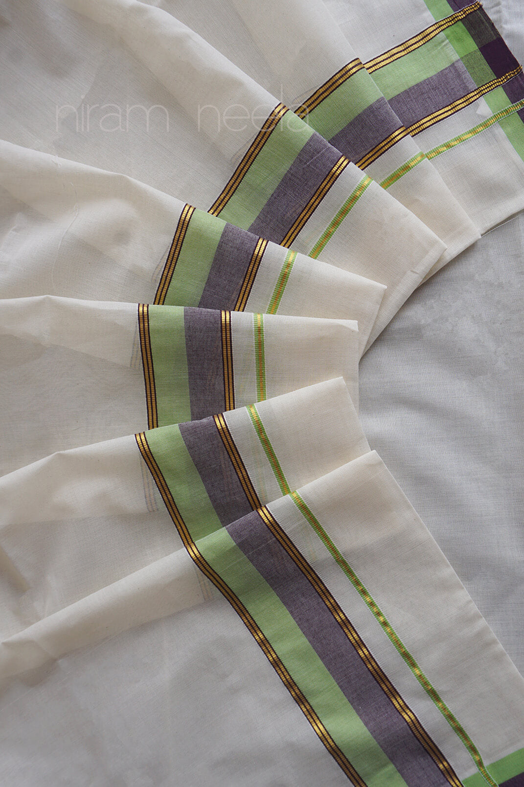A close-up of a white set mundu, featuring a border with stripes of light green, lavender-purple, and thin gold metallic lines.