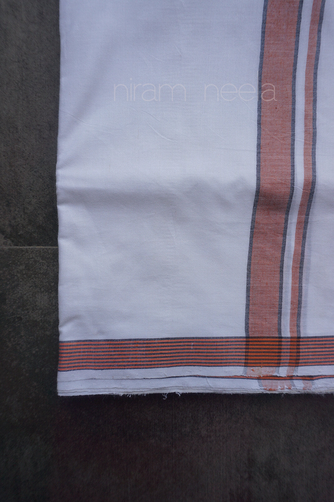 A close-up of a white cotton dhoti, featuring a vertical rust-orange stripe with thin white pinstripes, and a matching horizontal striped border. The fabric is laid flat on a dark gray surface, with a faint watermark 'niram neela' visible in the center.
