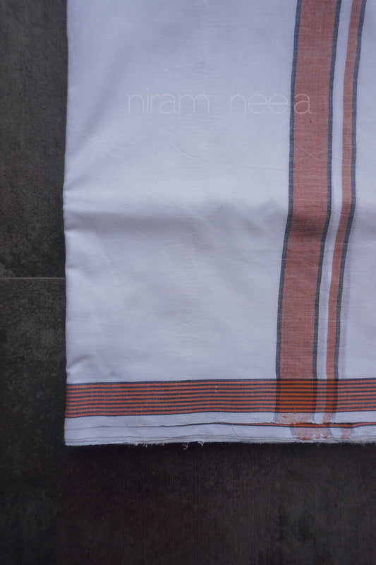 A close-up of a white cotton dhoti, featuring a vertical rust-orange stripe with thin white pinstripes, and a matching horizontal striped border. The fabric is laid flat on a dark gray surface, with a faint watermark 'niram neela' visible in the center.