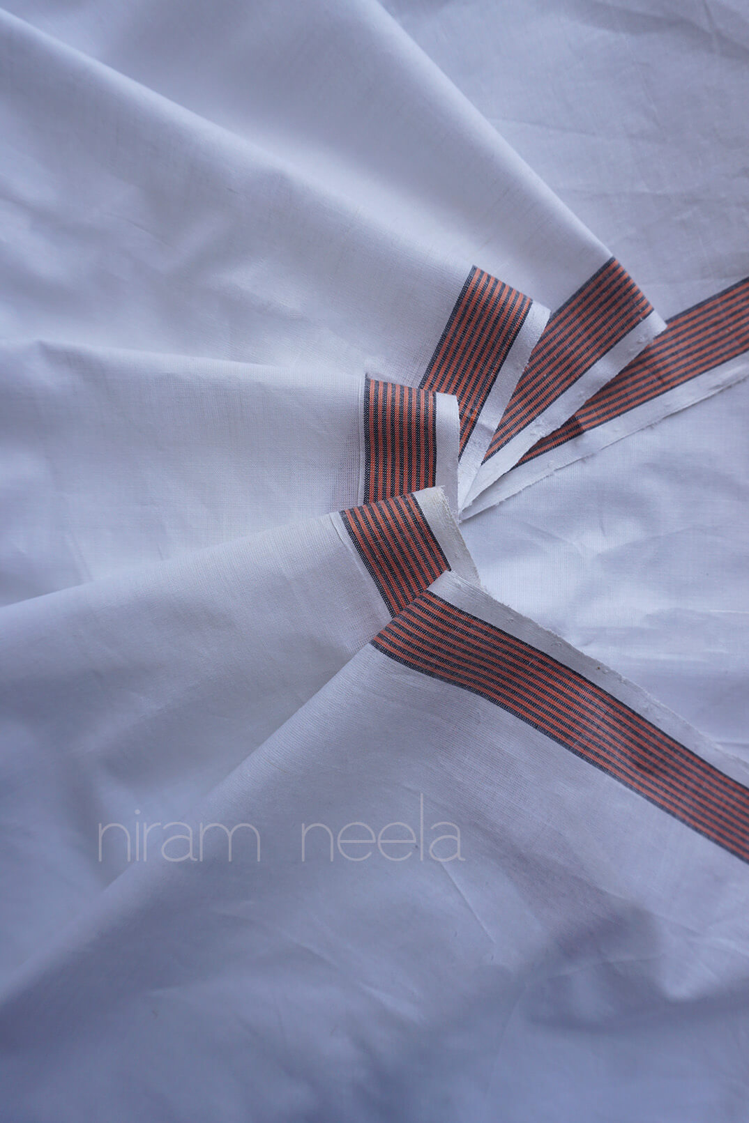 White dhoti with narrow reddish-brown and orange striped borders radiating from a central point, shown in a close-up studio-style shot with a faint 'niram neela' watermark.