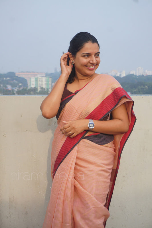 Woman wearing a peach saree with a red border, standing outdoors with a blurred background.