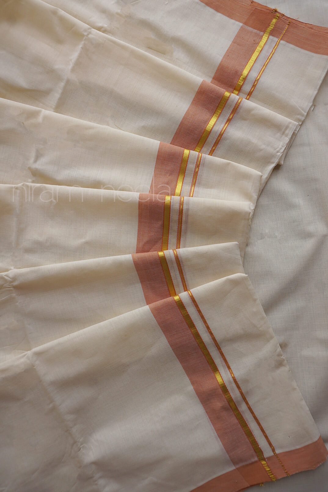 A close-up of a cream-colored mundu, featuring a peach-pink border with thin gold stripes running down the center.