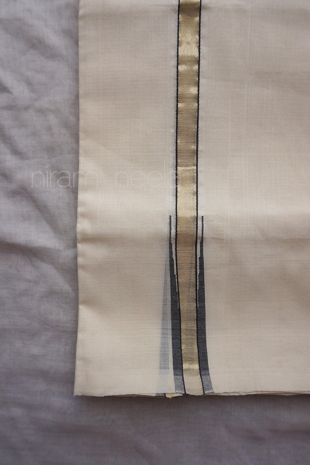 A close-up of a cream-colored dhoti, featuring a vertical metallic gold stripe flanked by thin black stripes, laid flat on a gray textured background.