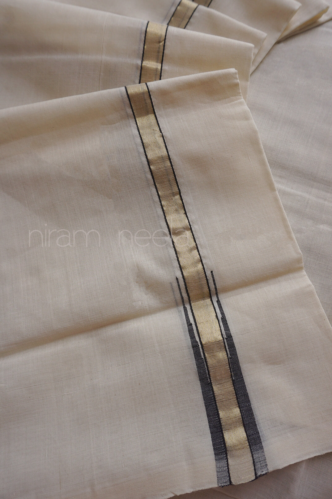 A close-up of a folded cream-colored dhoti in cotton, featuring a central vertical stripe pattern with a metallic gold center and thin black side borders.