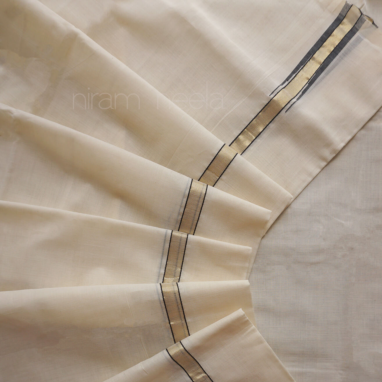 A close-up of a cream-colored dhoti, with a subtle sheen and a decorative border featuring black and gold stripes. The fabric is arranged in soft, radiating folds.