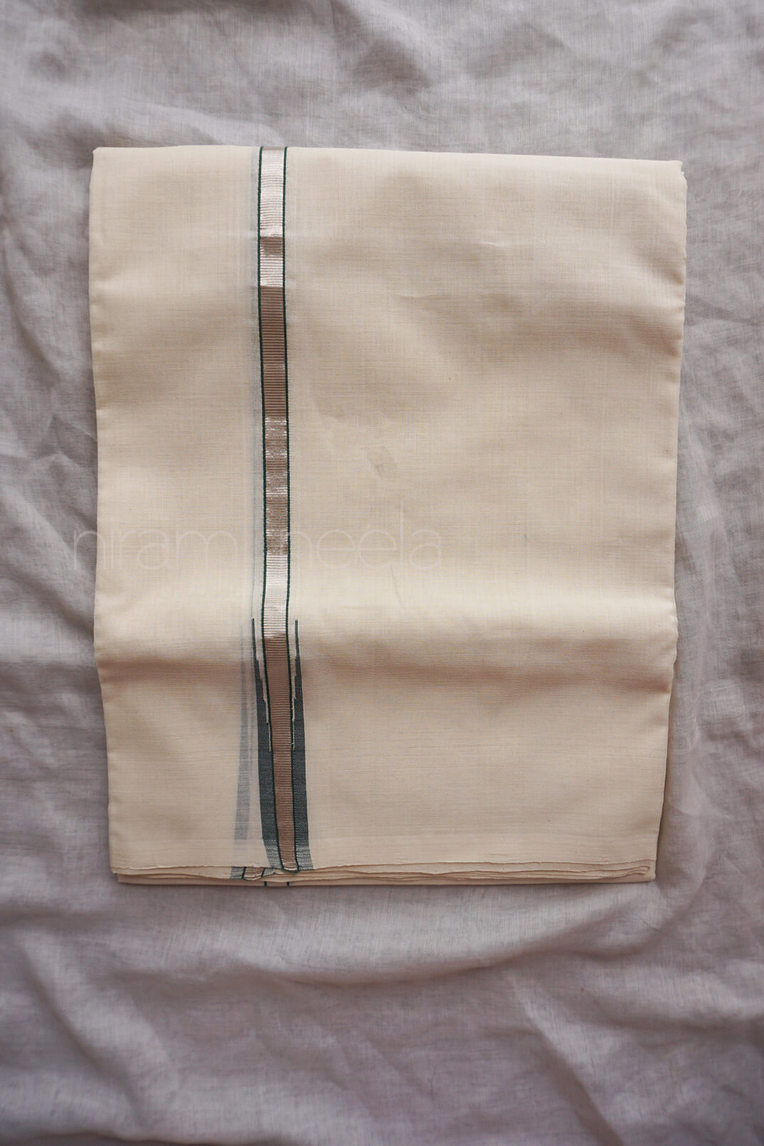 A folded cream-colored cotton dhoti,  featuring a vertical green silver stripe, laid flat on a textured light gray fabric background.