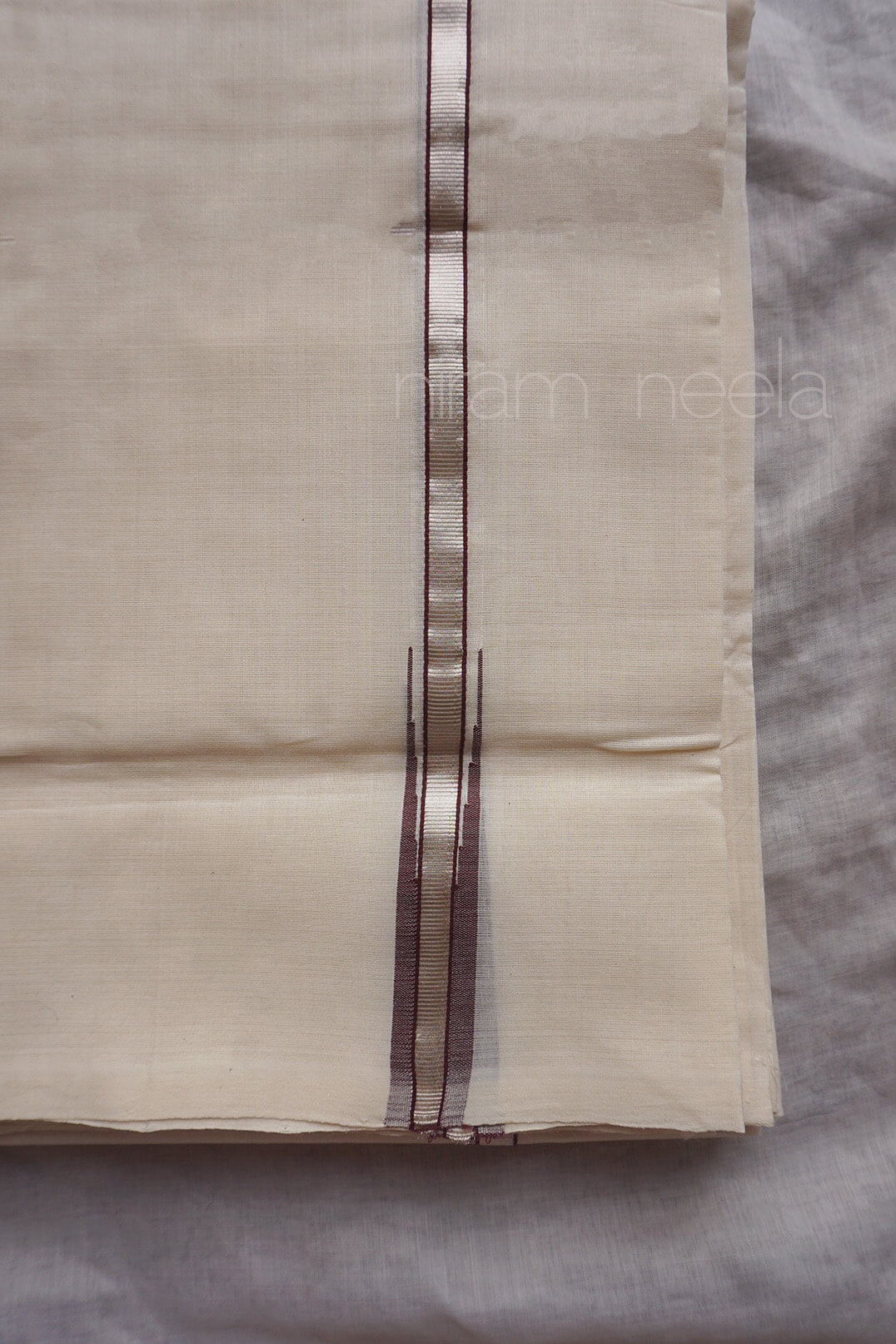 A folded cream-colored cotton dhoti,  featuring a vertical metallic brown border stripe down the center, laid flat on a gray textured surface.